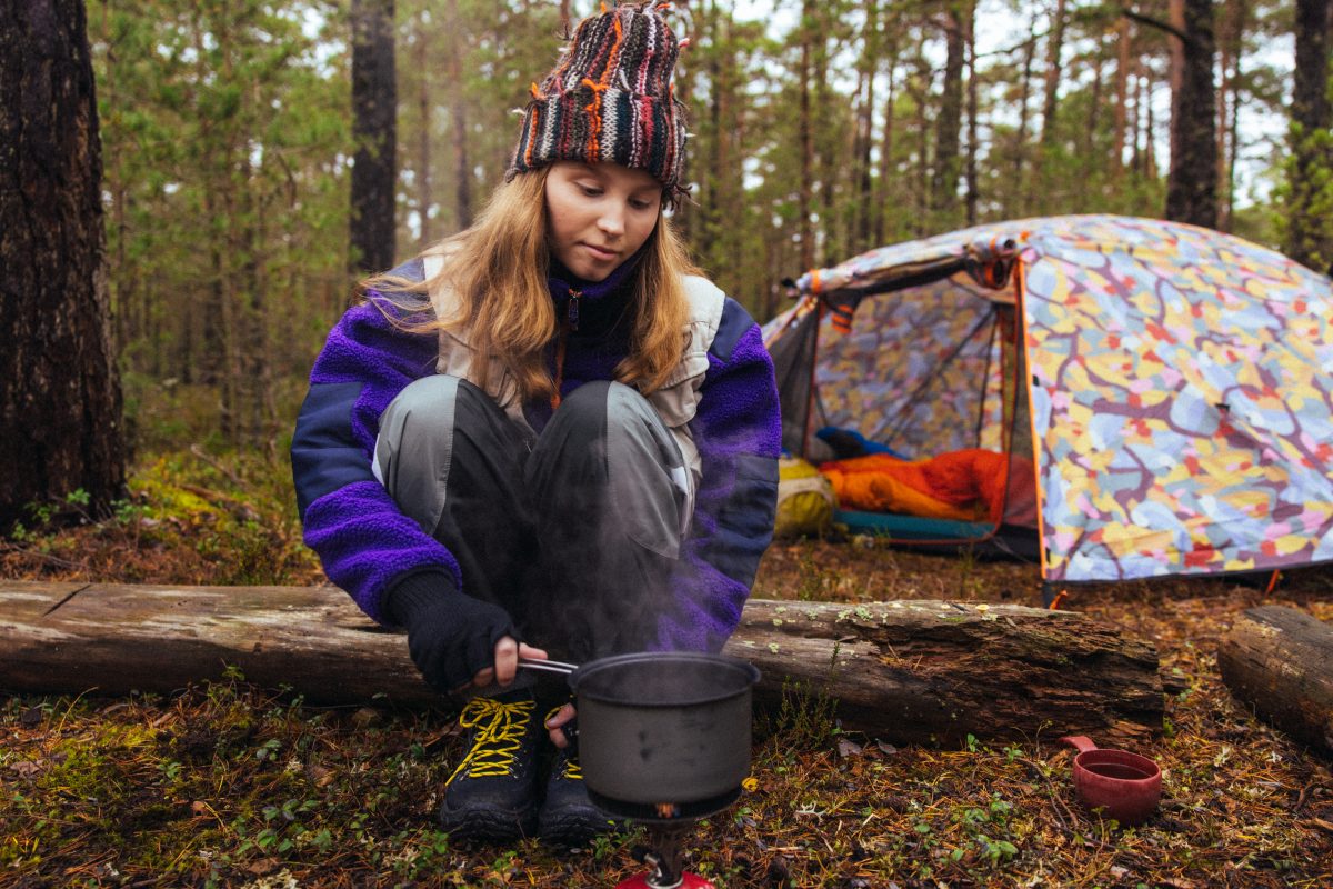 Nuori retkeilijä istuu metsässä keittimen äärellä valmistamassa ruokaa. Taustalla näkyy värikäs teltta, jonka sisällä on makuupussi ja varusteita. Metsän vehreä maasto ympäröi tunnelmallista retkeilytilannetta.